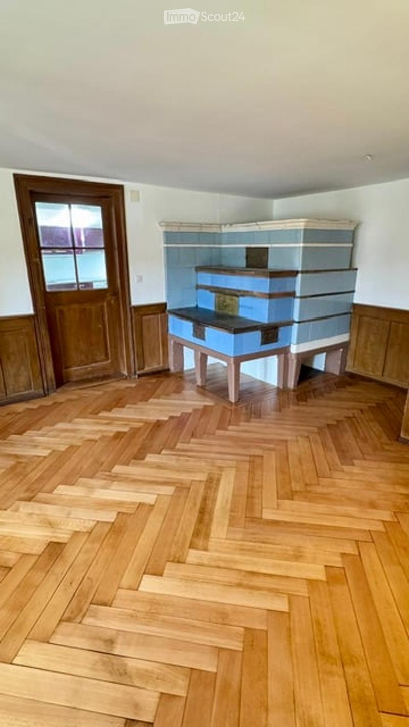Empty room with wooden herringbone flooring, white walls, and a blue fireplace. The room features a wooden door and a window with a view of the outside.