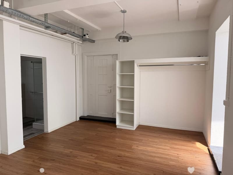 Empty room with wooden flooring, white walls, a built-in closet with shelves, a hanging light fixture, and a doorway leading to a tiled bathroom area.