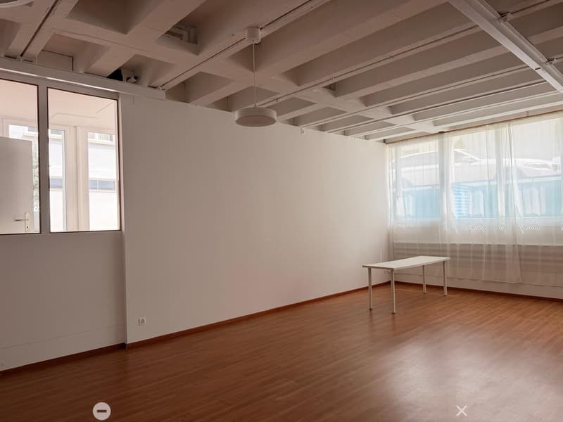 Empty room with wooden flooring, white walls, large windows with sheer curtains, and a single small table near the window.
