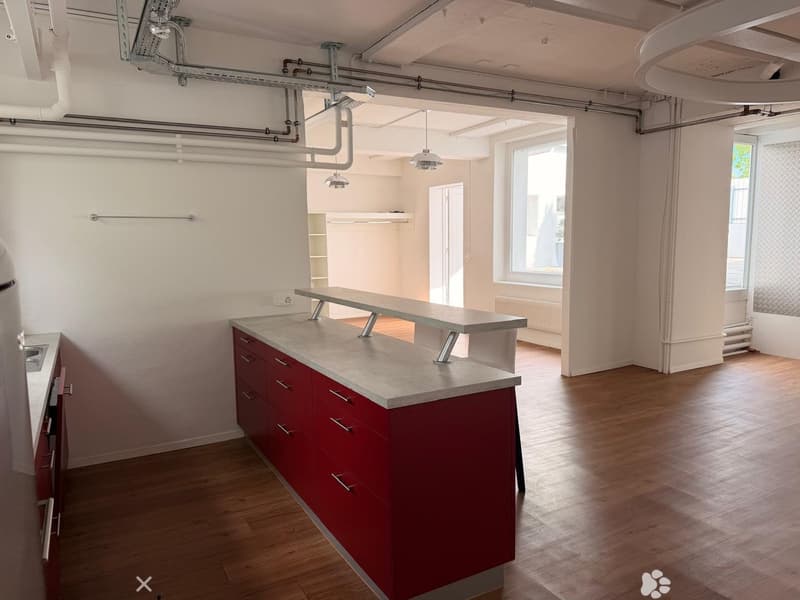 Modern kitchen with red cabinets, a central island, and wooden flooring. Exposed pipes and lighting fixtures are visible on the ceiling.