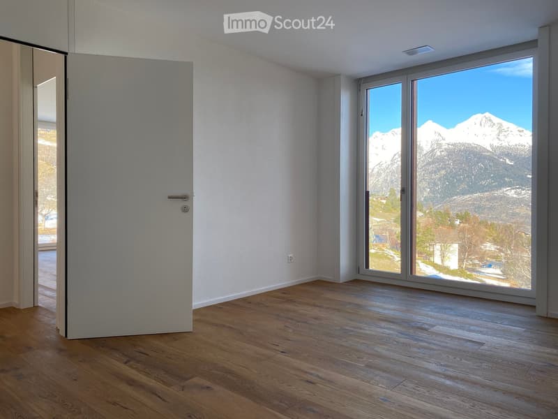 Empty room with wooden flooring, large windows offering a mountain view, and a closed door.