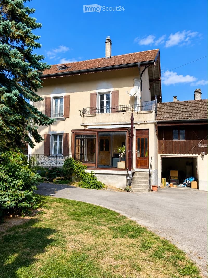 Two-story house with beige walls, brown shutters, and a tiled roof. Features include a garage, a small balcony, and a well-maintained lawn.