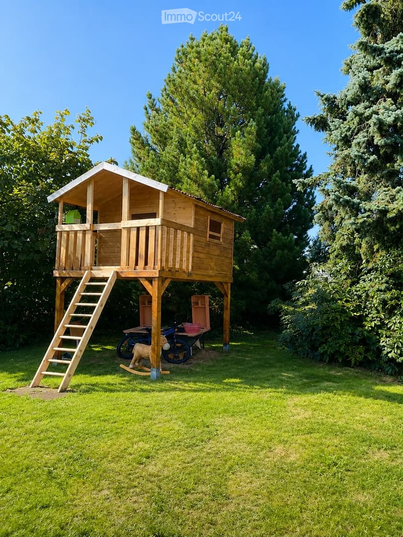 A wooden treehouse with a staircase, surrounded by lush green trees and grass. The treehouse has a balcony and a small window, with a toy horse and a wheelbarrow nearby.