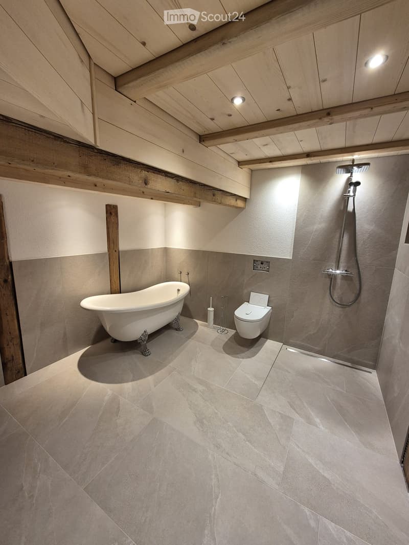 Modern bathroom with a freestanding bathtub, toilet, and shower, featuring exposed wooden beams and recessed lighting.