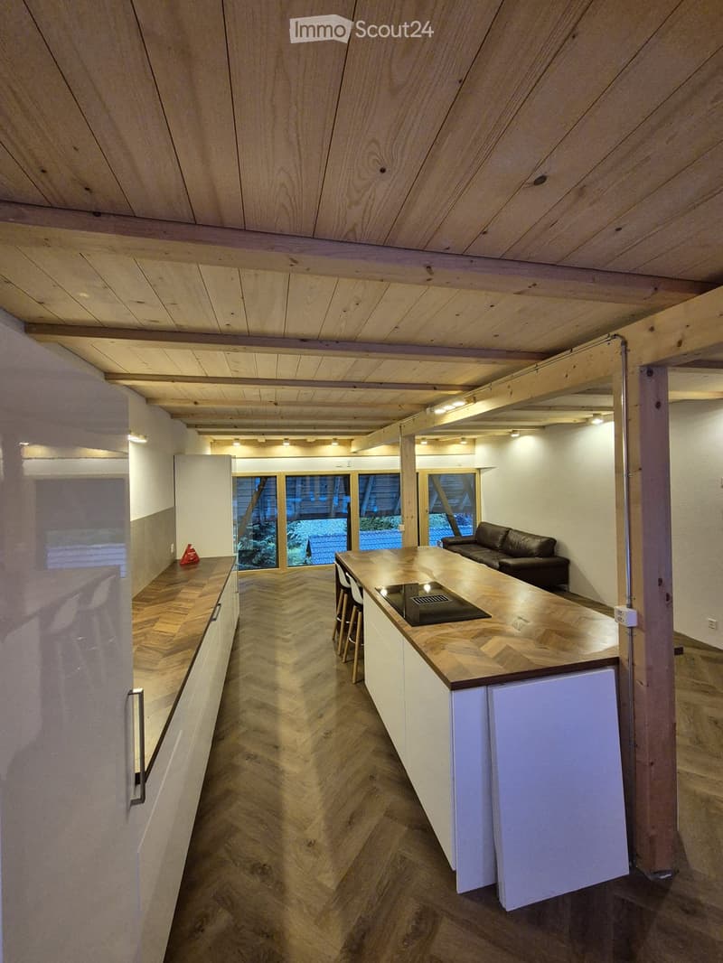 Open-plan living and kitchen area with wooden herringbone flooring, white kitchen cabinets, a central island with a countertop, and a black leather sofa. Large windows provide natural light and a view of the outdoor area.