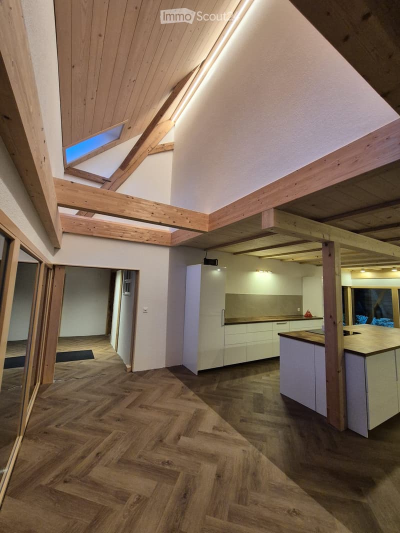 Spacious, well-lit room with exposed wooden beams and herringbone wooden flooring. Features modern kitchen cabinets, a refrigerator, and a countertop.