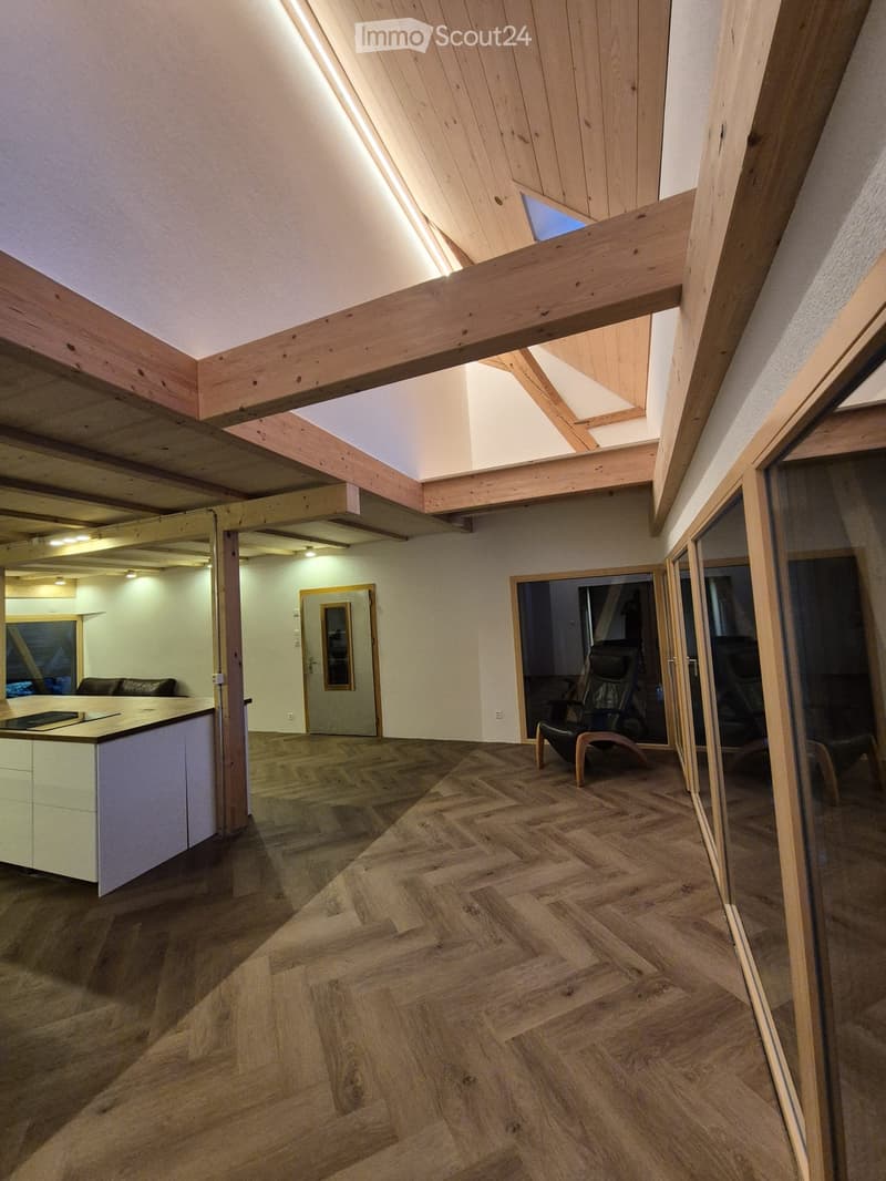 Bright, open living space with wooden herringbone flooring, exposed wooden beams, and large windows. Features a modern kitchen area with white cabinets, a sofa, and a comfortable chair near the window.