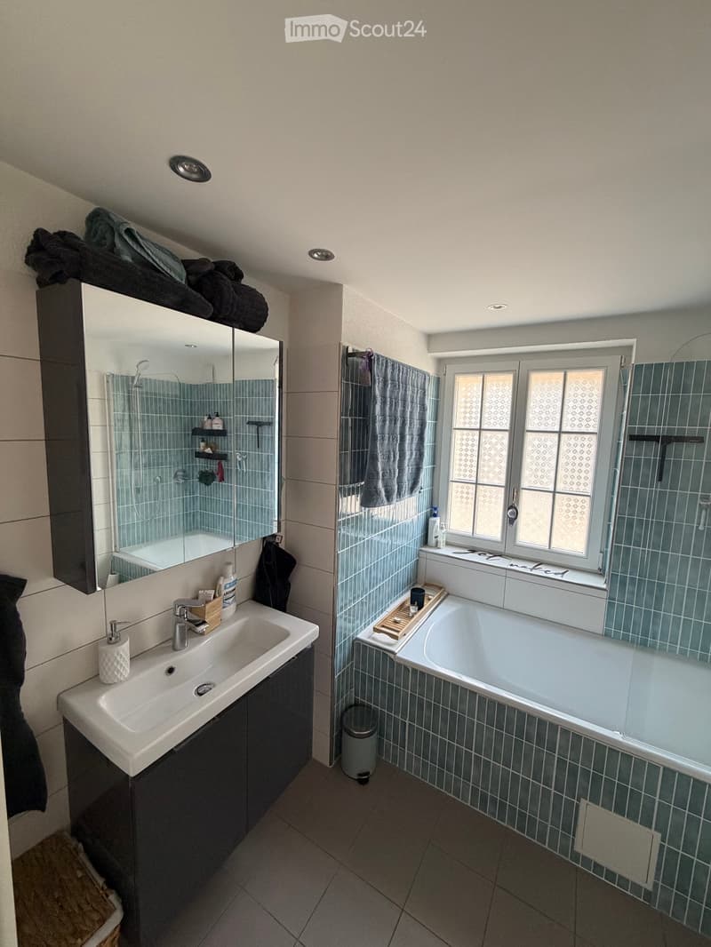 Modern bathroom with a large sink, bathtub, shower, and mirrored cabinet. Features include a window with frosted glass, towels, and toiletries.