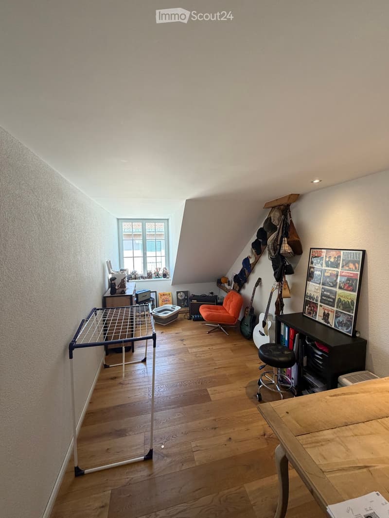 A cozy attic room with wooden flooring, slanted ceilings, and a large window. Contains a table, chair, guitar, stool, shelf with books and posters, cat litter box, and a bed frame.