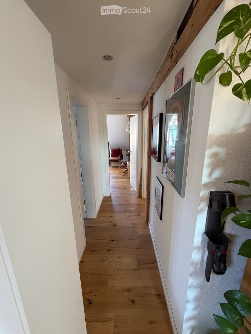 A narrow hallway with wooden flooring, white walls, and several framed pictures on the right side. A potted plant is visible on the right, and a doorway leads to another room with a red chair and a small table visible in the background.