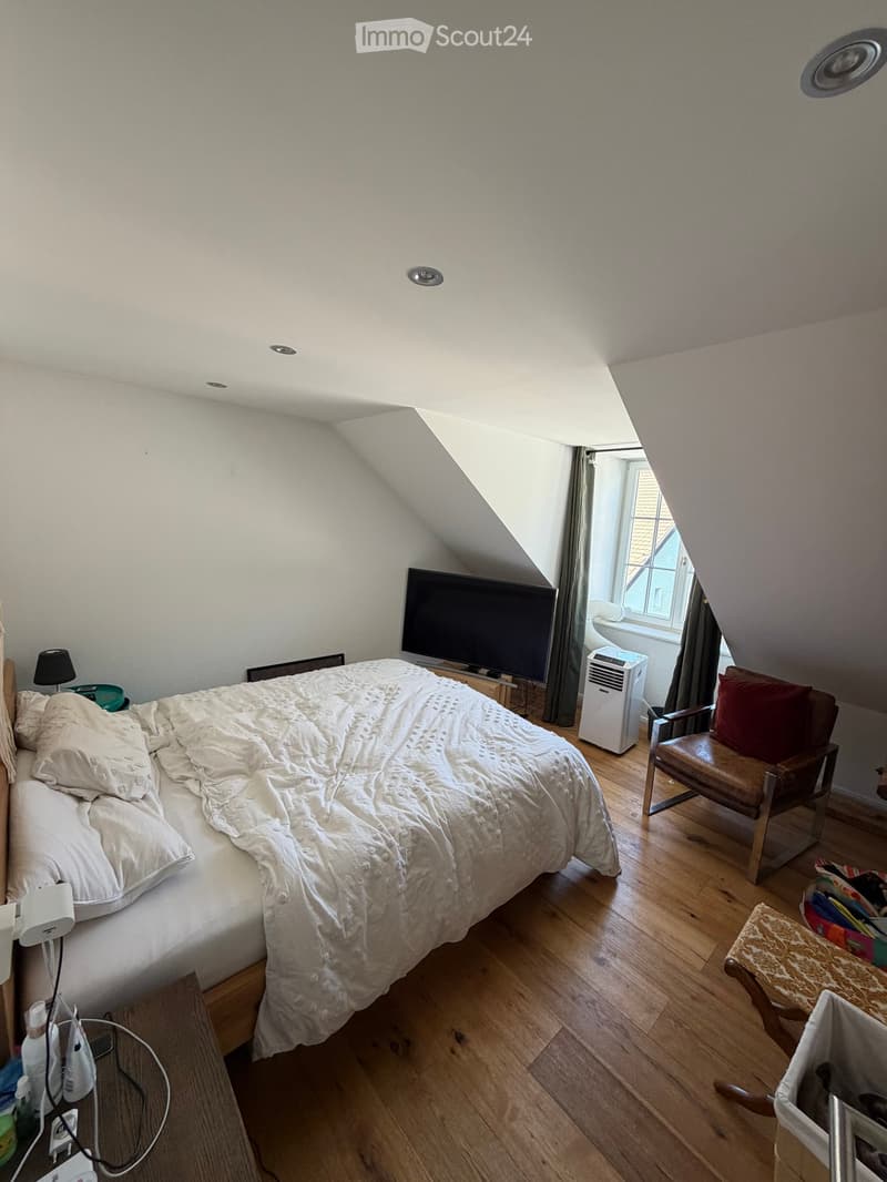 Cozy attic bedroom with wooden flooring, a large bed with white bedding, a wooden chair with a red cushion, a small round table, a television, and an air conditioner unit near the window.