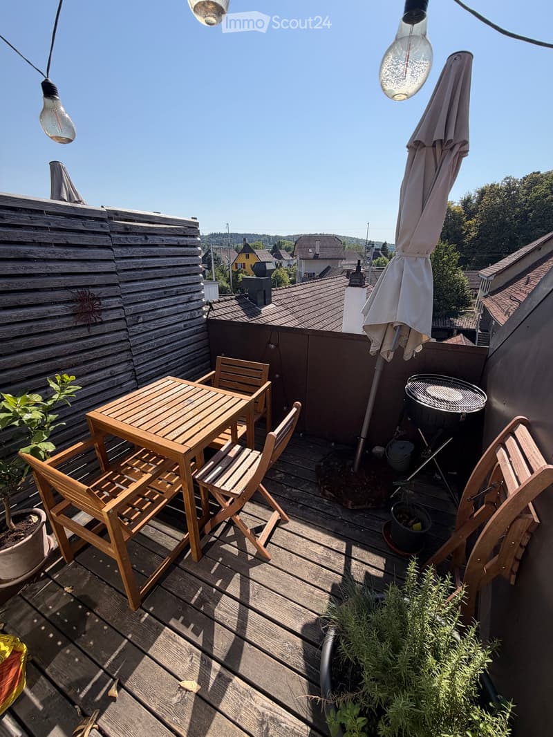 Wooden terrace with a small table, chairs, umbrella, grill, and potted plants, overlooking a residential area.