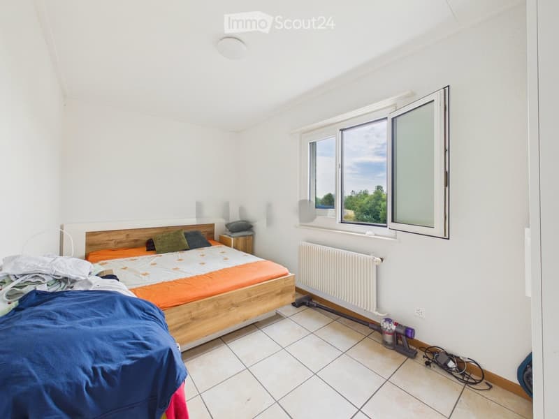 Bright bedroom with a wooden bed, orange and blue bedding, nightstand, radiator, and a vacuum cleaner on the floor near the window.