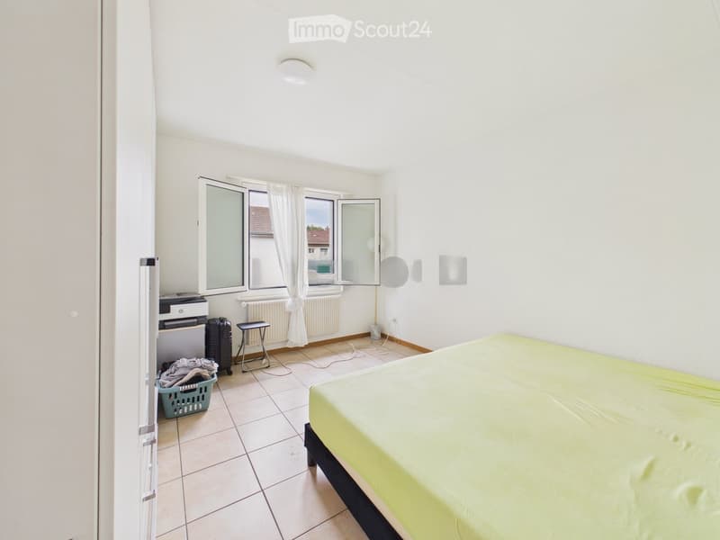 Bright bedroom with a single bed, a small stool, a printer, a suitcase, and a laundry basket near the window.