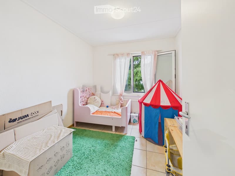A small, well-lit bedroom with a single bed, a colorful toy tent, a small table, and a chair. The room features a green rug and large windows with white curtains.