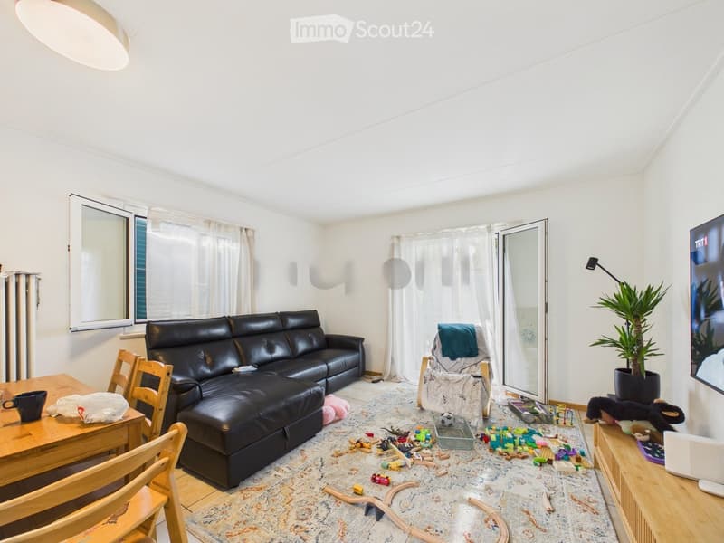 Living room with a black leather sofa, wooden dining table, chairs, TV, and scattered toys on the floor.