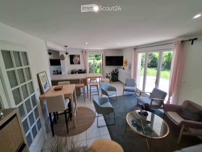 Modern living room with a sofa, coffee table, chairs, a dining table with stools, and a TV screen. Large windows and sliding doors lead to a garden area.