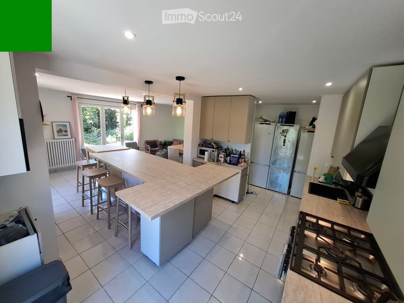 Modern kitchen with a central island, bar stools, and various appliances including a refrigerator, microwave, and oven. The space features light-colored cabinets, a tiled floor, and pendant lights.