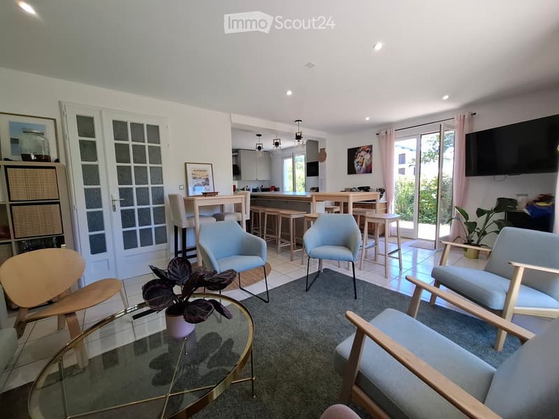 Modern living room with a glass coffee table, light blue chairs, a grey rug, and a kitchen area with bar stools visible in the background.