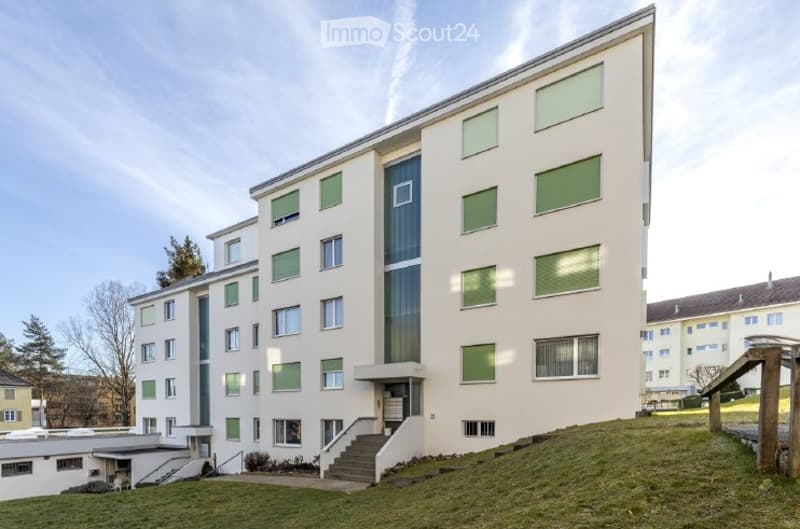 Modern multi-story residential building with light-colored facade, green-framed windows, and a raised ground floor entrance.