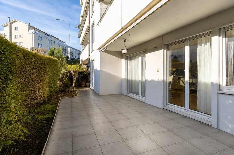 Balcony with tiled floor, large glass doors leading to a room, white walls, and a small garden area with bushes and plants.