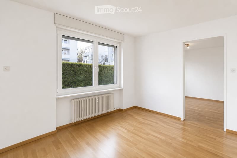 Empty room with wooden flooring, white walls, large windows with blinds, radiator, and visible electrical outlets.