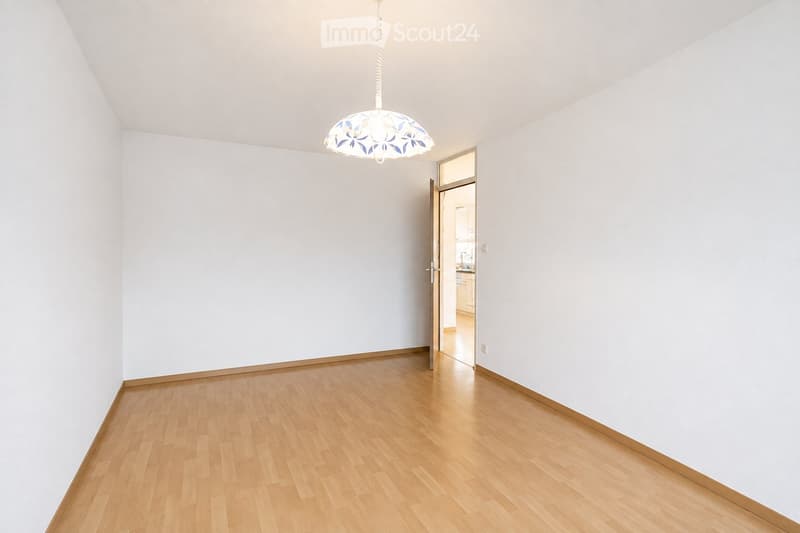 Empty room with light wooden flooring, white walls, and a hanging light fixture. A doorway leads to another room.