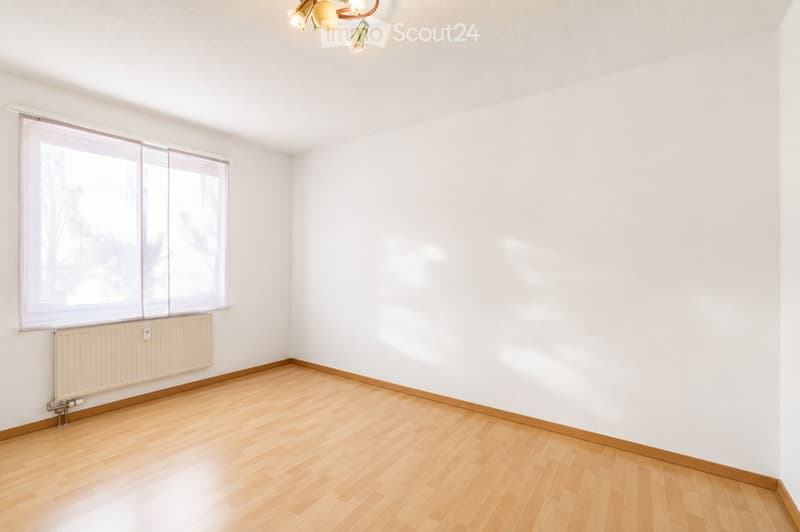 Empty room with light wooden flooring, white walls, large window with blinds, radiator, and a ceiling light fixture.