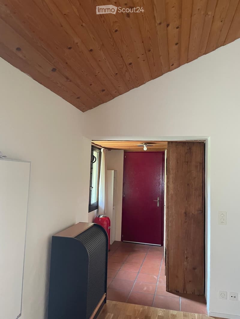 A narrow hallway with wooden ceiling beams, white walls, and a red door leading to another room. The floor is a mix of wooden planks and tiled sections.