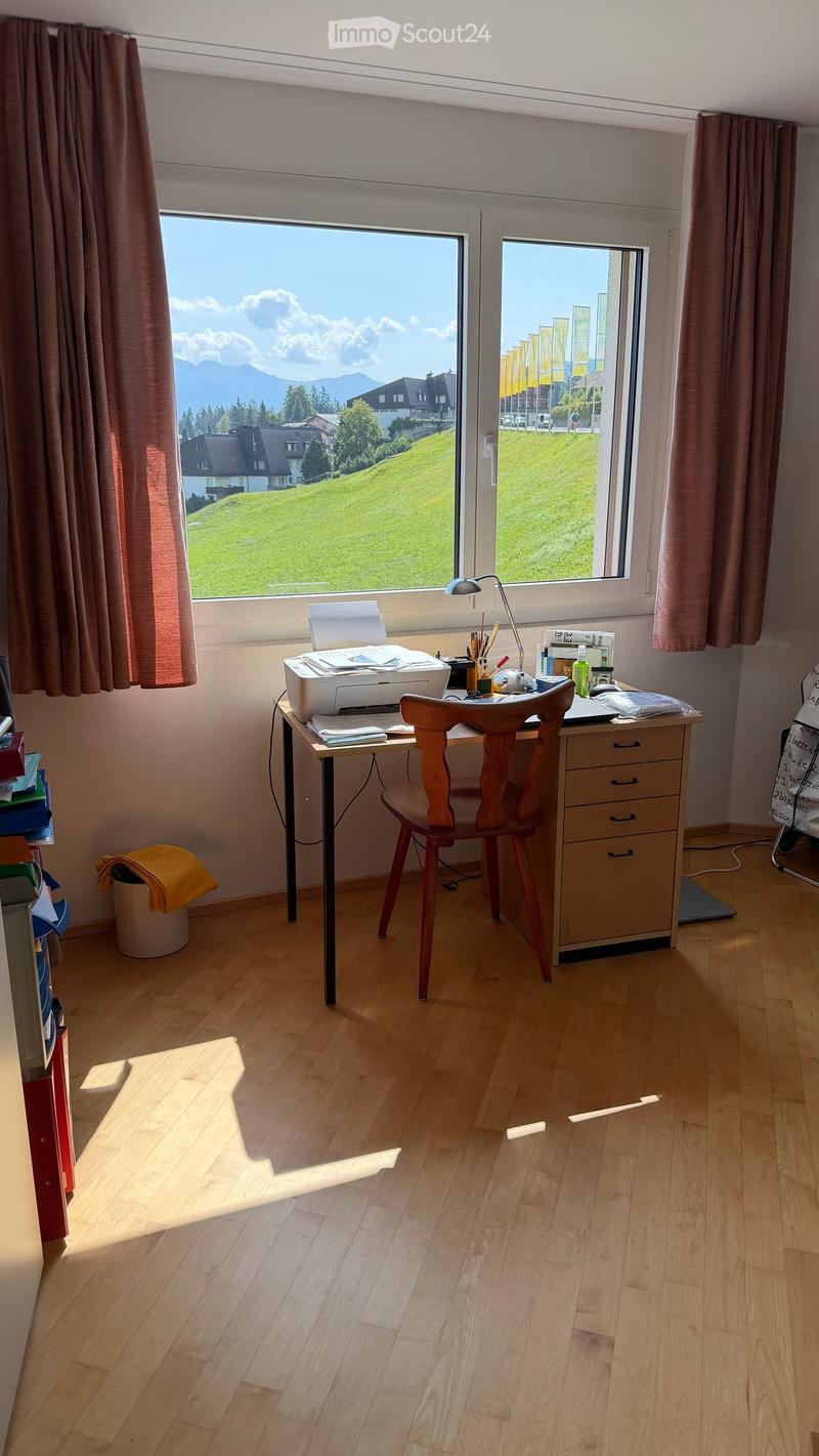 Brightly lit room with wooden flooring, large window offering a scenic view of a green field and mountains, a desk with a printer, chair, and various office supplies, and a wooden cabinet.