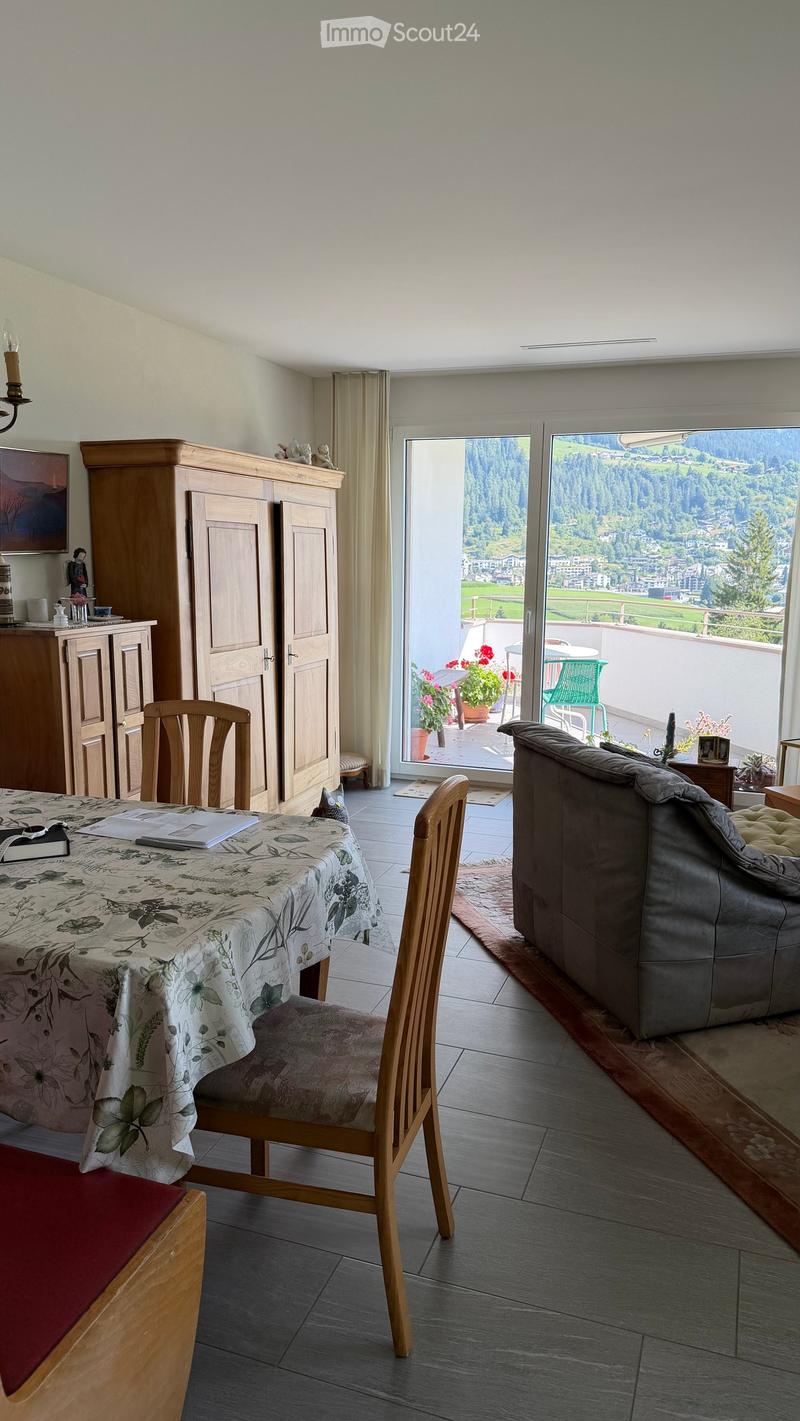Bright living room with a view of the mountains, featuring a sofa, dining table with chairs, wooden cabinet, and large windows with curtains.