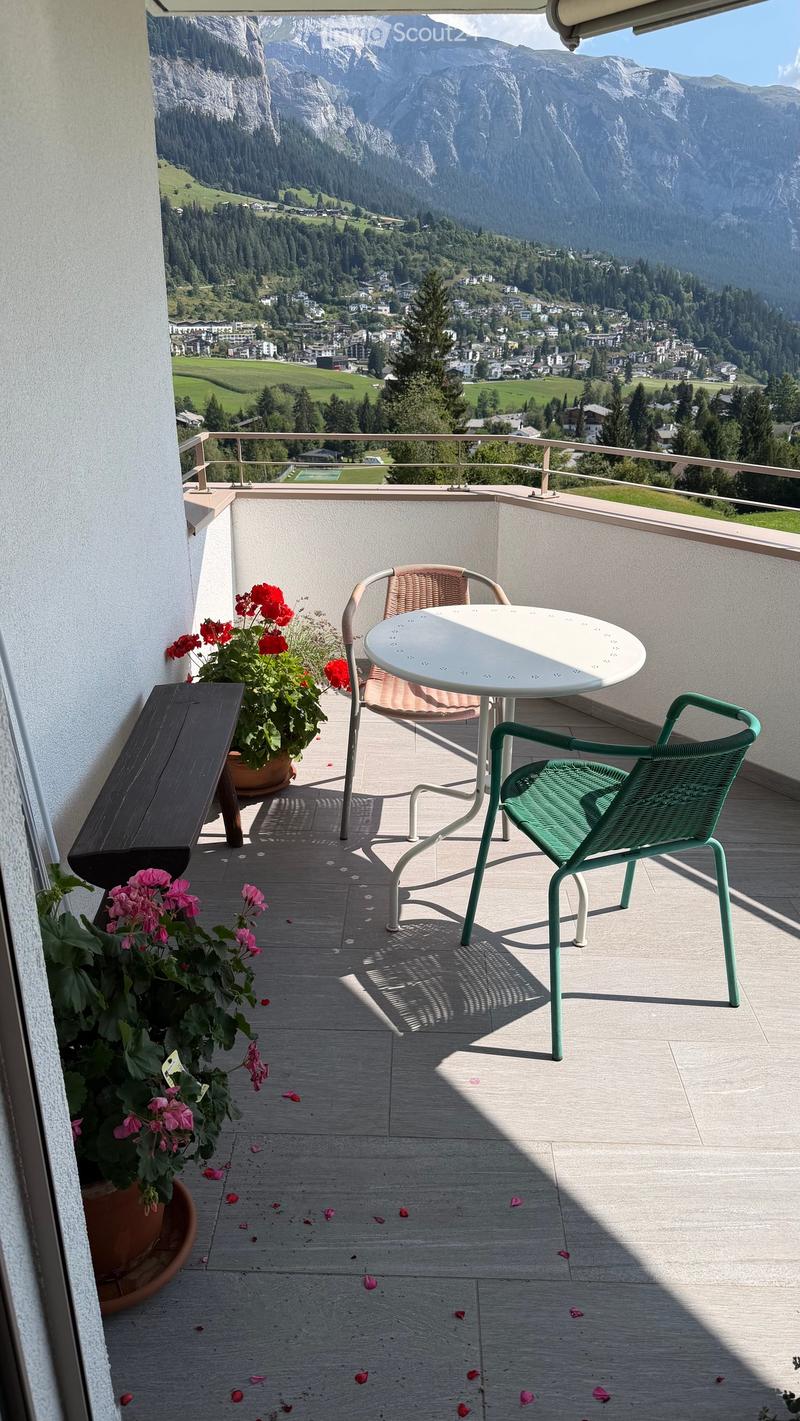 Balcony with a scenic mountain view, featuring a round white table, green wicker chair, wooden bench, and two flower pots with red and pink flowers.