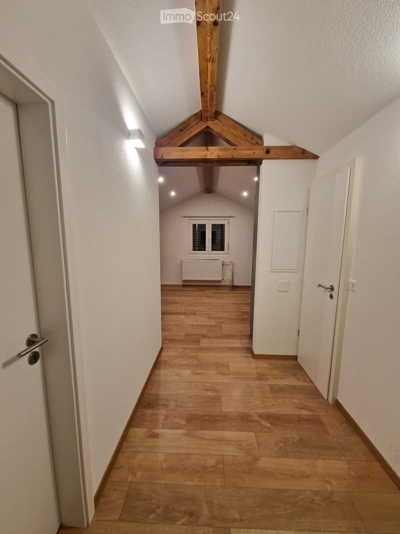 Spacious hallway with wooden floor and white walls, wooden beam ceiling, white doors, mounted light, and small window.