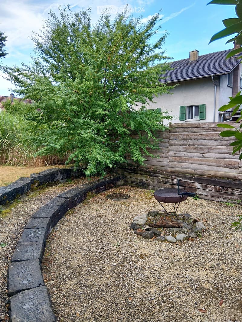 Garden area with gravel ground, stone border, metal fire pit, and wooden fence. A house with green shutters is visible in the background.
