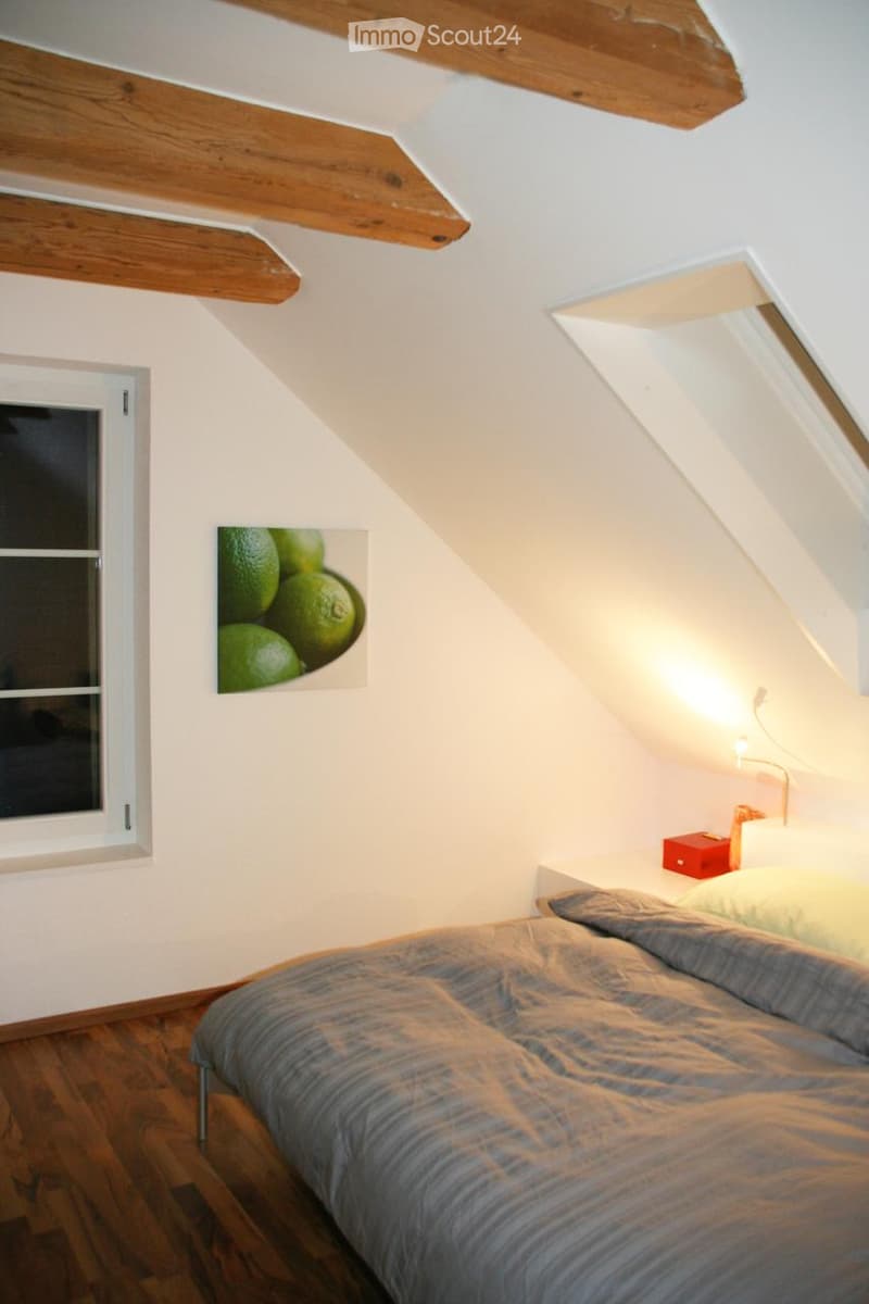 Cozy attic bedroom with wooden slanted ceilings, a bed with grey bedding, a nightstand with a lamp, and a picture of limes on the wall.