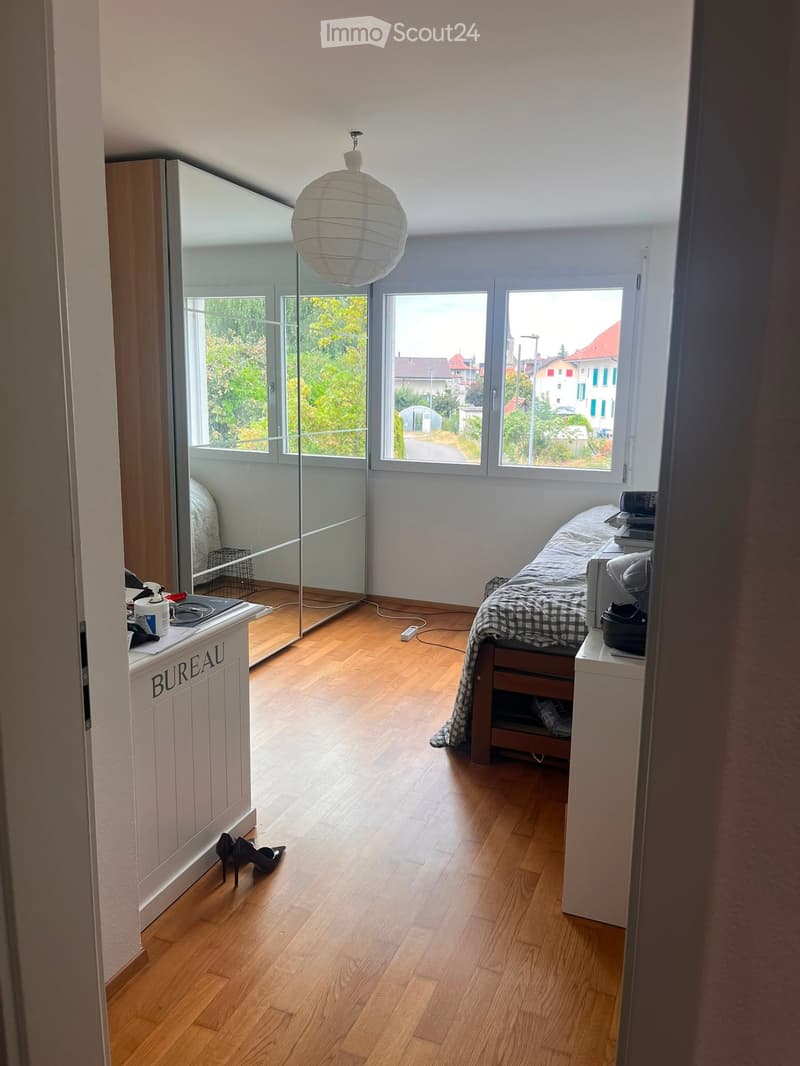 Bright bedroom with wooden flooring, large windows, a bed with a patterned blanket, a nightstand, a mirror, and a hanging lamp.