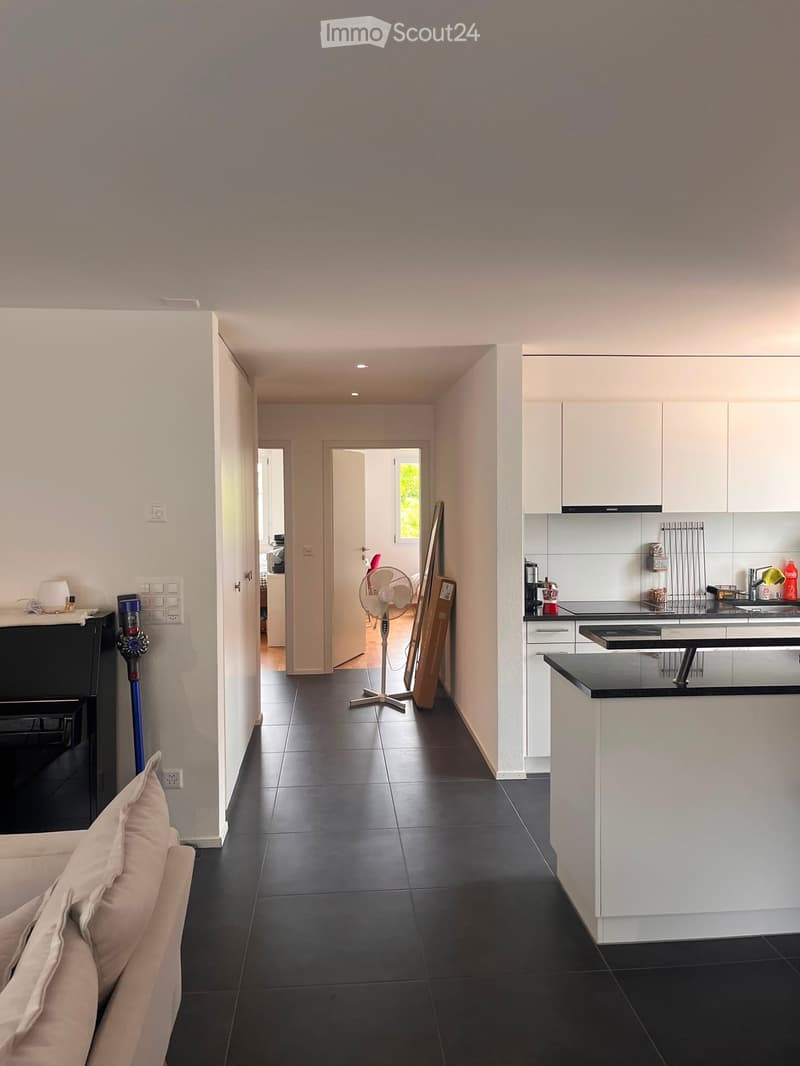 Open-plan living and kitchen area with dark tiled flooring, white kitchen cabinets, a sofa, a standing fan, and a small table with chairs.