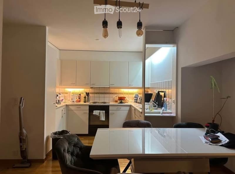 A modern, open-plan kitchen and dining area with white cabinets, a central island, and a dining table with chairs. The kitchen features an oven, fridge, and sink, while the dining area includes a table and chairs. A vase with a plant is placed near the wi