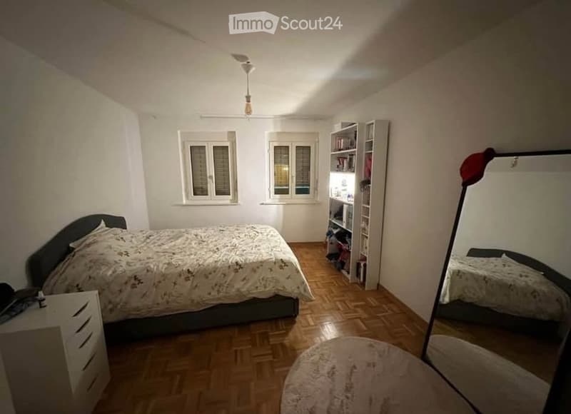 Cozy bedroom with wooden flooring, a bed with a patterned duvet, a nightstand, a large mirror, and a bookshelf filled with books.
