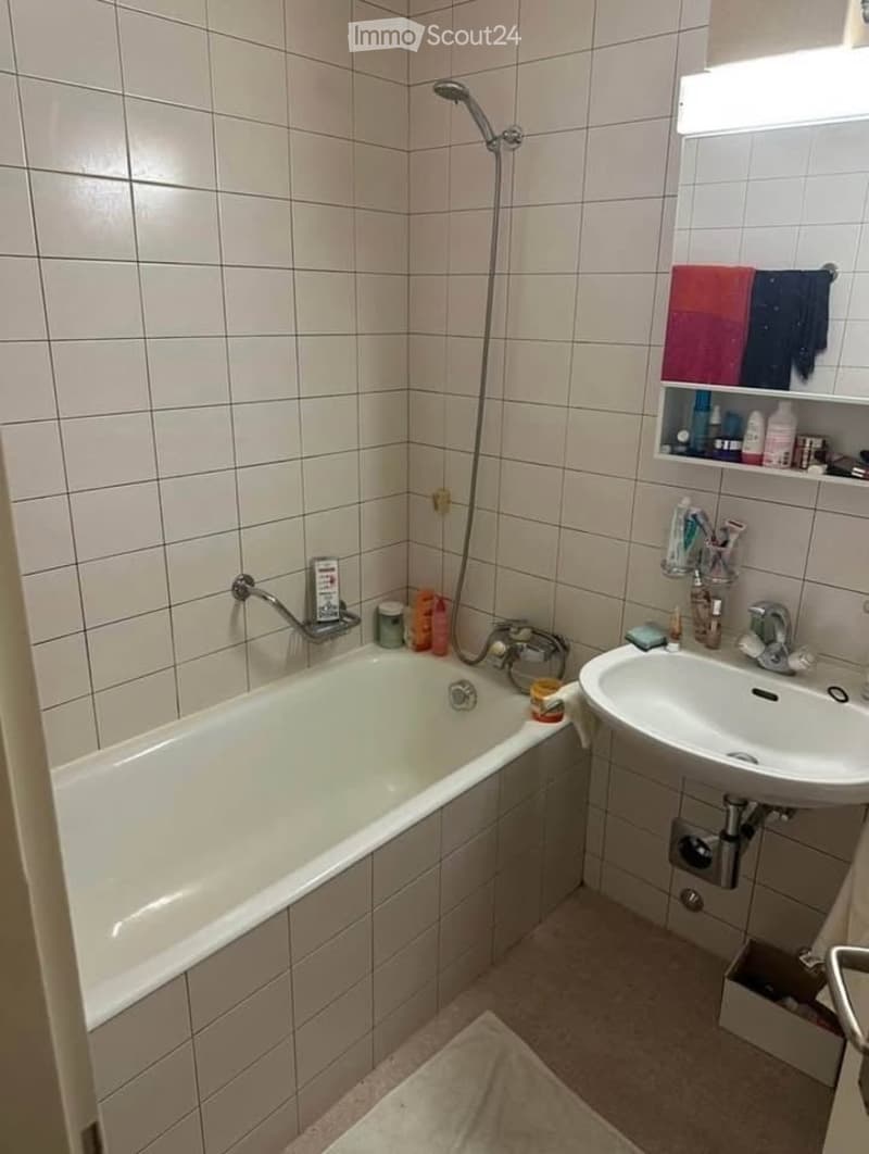 A small bathroom with a white tiled wall and floor, featuring a bathtub, a sink with a faucet, a showerhead, a towel on the floor, and a shelf with toiletries.