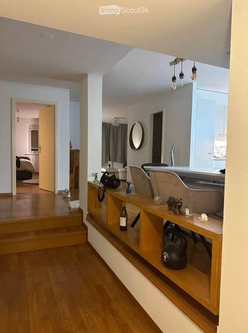 Modern open-plan living and dining area with wooden flooring, built-in shelving, a sofa, chairs, a dining table, and decorative items including a mirror and a bottle on the shelf.