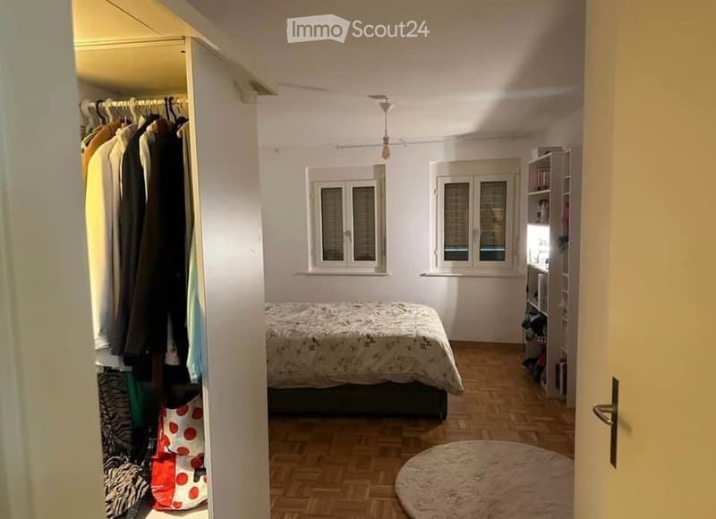 Cozy bedroom with wooden flooring, a bed with a patterned duvet, a wardrobe with hanging clothes, a bookshelf, and a round rug on the floor.