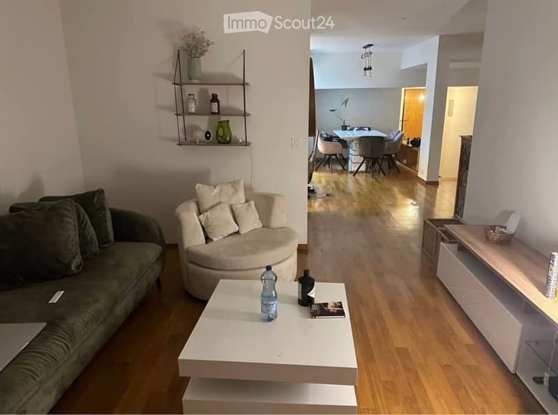 Modern living room with a green sofa, white coffee table, round chair, and shelves. The room features wooden flooring and opens into a dining area with a table and chairs.