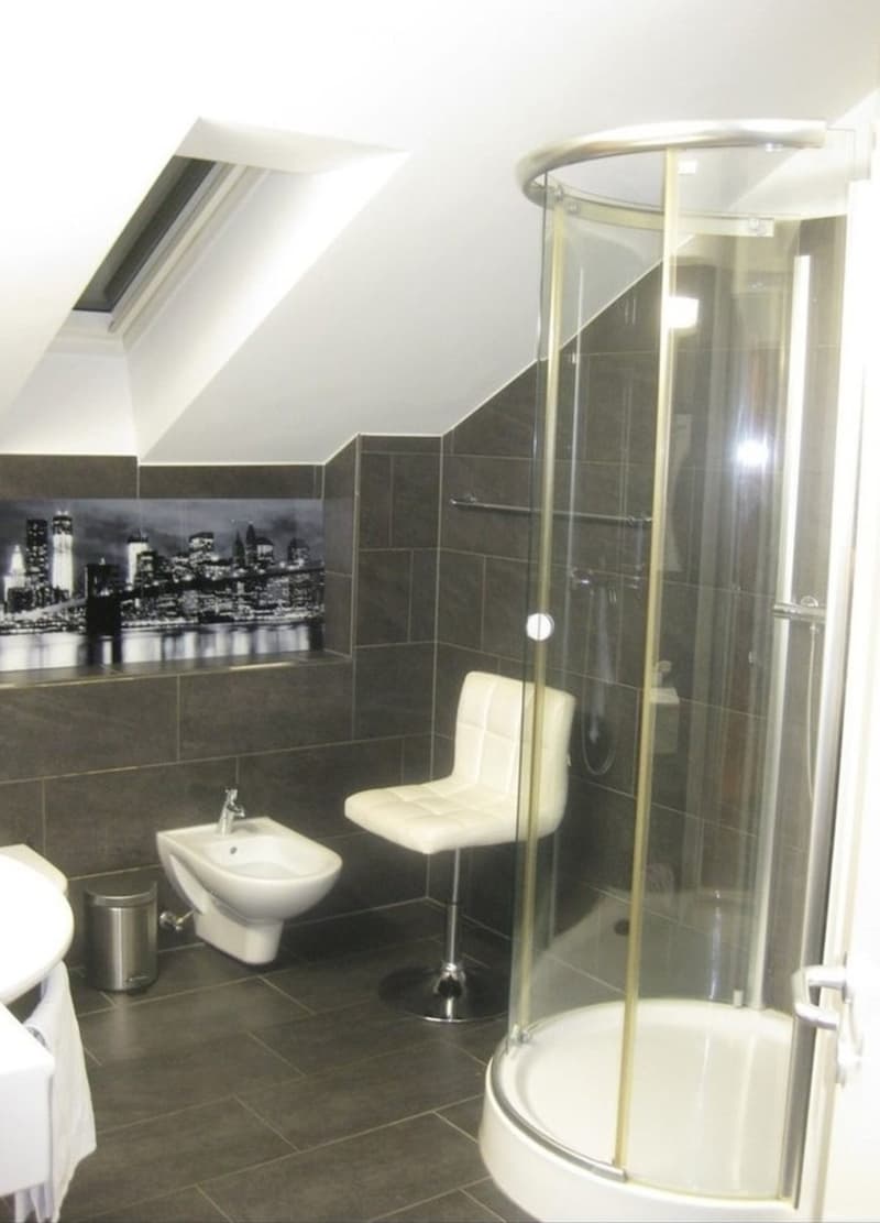 Modern bathroom with dark tiles, a glass-enclosed shower, a white stool, a toilet, and a sink.