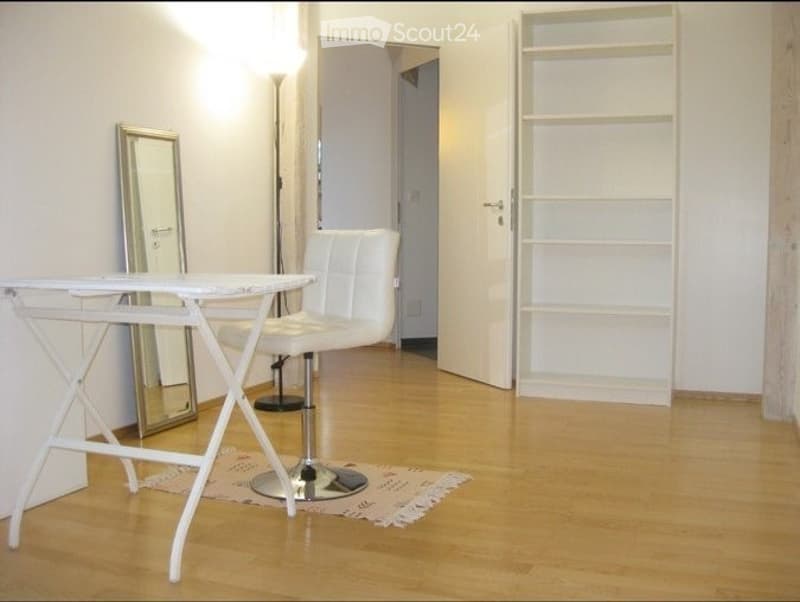 A modern, minimalist room with a white leather chair, a small white table, a mirror, and a white shelf unit. The floor is light wood, and the walls are white.