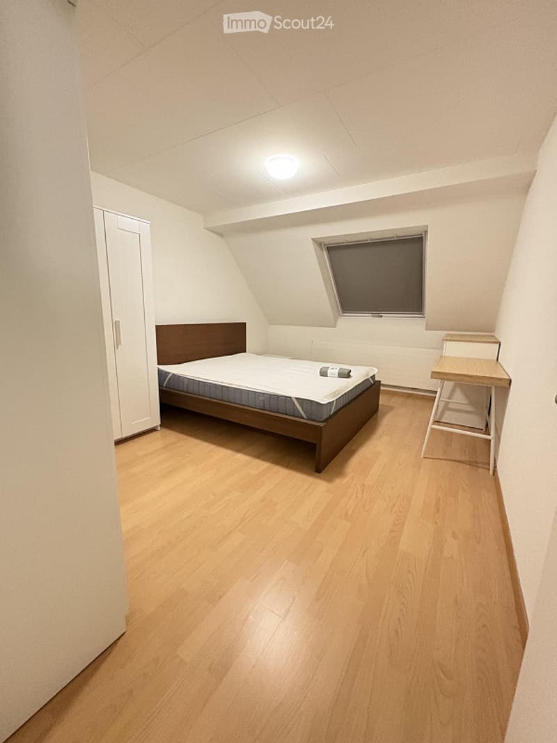 Cozy bedroom with wooden flooring, a bed with a mattress, a nightstand, a small table, and a chair. The room features white walls and a slanted ceiling with a window.