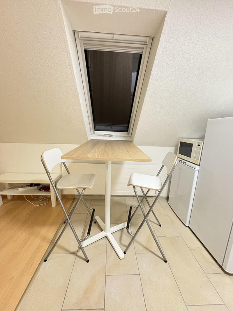 A small, furnished room with a wooden table, two white chairs, a refrigerator, and a microwave. The room has light-colored walls and a large window with blinds.