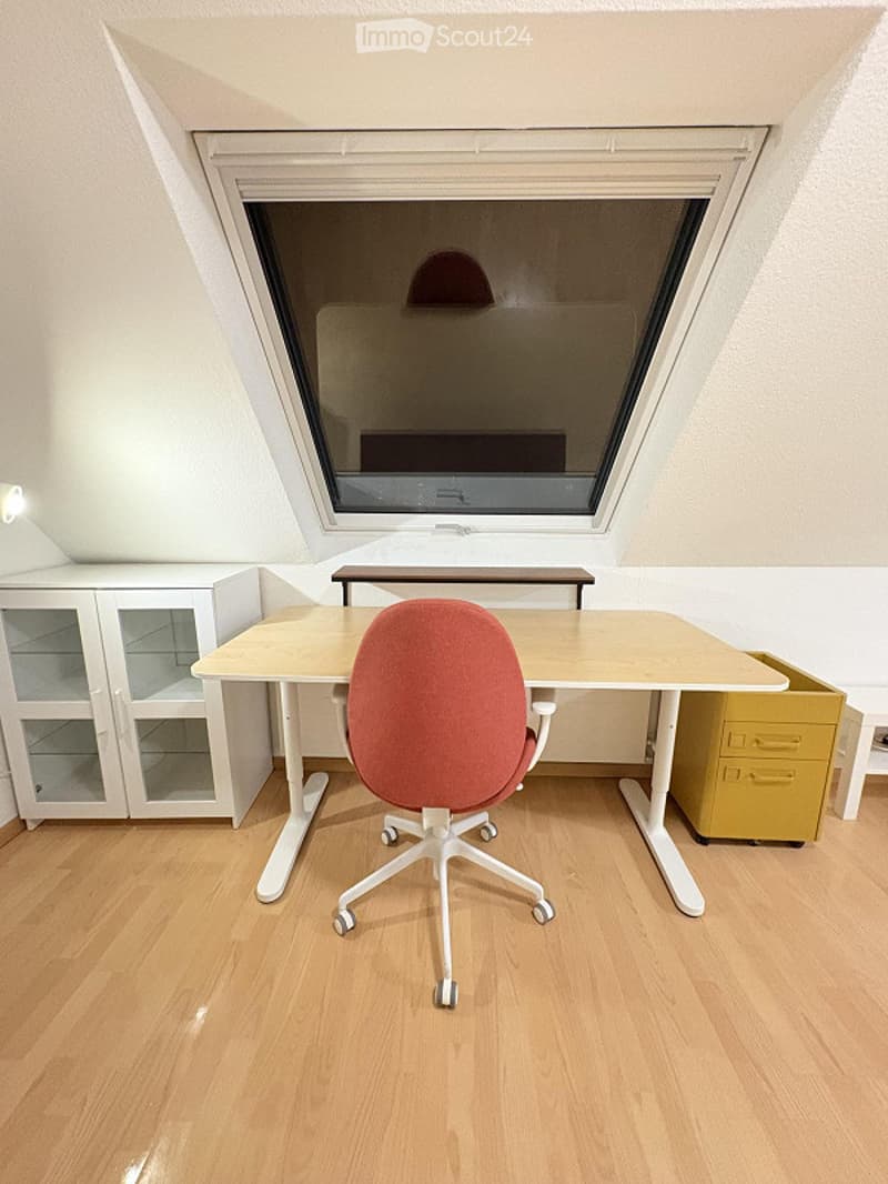 A modern, well-lit room with a slanted ceiling and skylight. Features a light wood desk, red office chair, white cabinet with glass doors, and a yellow drawer unit.