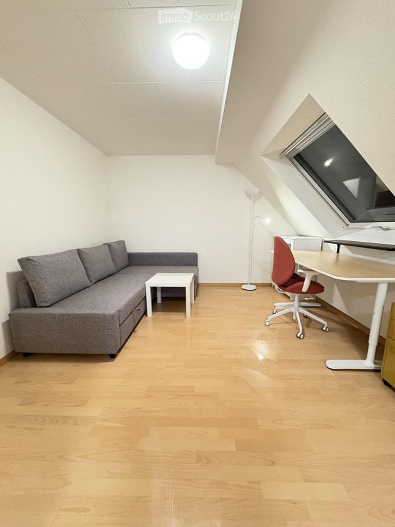 Modern, compact living space with a grey sectional sofa, small white coffee table, red office chair, white desk, and a floor lamp. The room features light wooden flooring and white walls, with a large window providing natural light.
