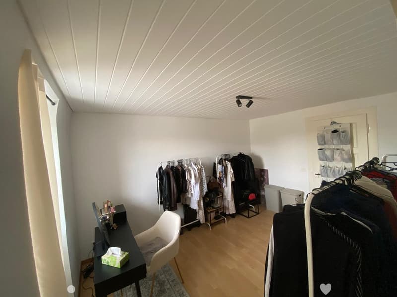 A well-lit room with wooden flooring, a white chair, a black table with a screen, and clothing racks with various garments.